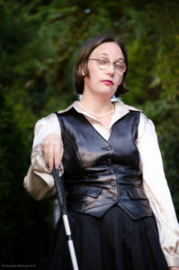 Author Elsa gazes down through her purple and gold vintage spectacles, wearing pink lipstick, a black leather vest, a white and gray striped silk shirt, and pearls. It’s unclear if she’s holding a sword or a white cane, but does it matter? 