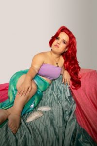 The author, Jillian, is lying on a sea-green ribbed blanket wearing a vibrant green and blue skirt reminiscent of Ariel's mermaid tail. They have scorchingly bright red hair and a purple tube top. They are leaning on their left side with the right hand laid on their right knee. Lying in front of them are two seashells and she is looking directly at the viewer with eyes tinted with red eyeshadow. They are lying as a noble princess would.