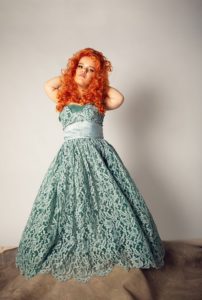 The author, Jillian, is standing in a stunning blue-green strapless dress with white lace and a sash-covered waist ribbon. They have bright orange curly hair that falls below their shoulders and a gaze that looks past the viewer. They are holding their hands up, clutching the back of their head while standing on a grey mound with lighter grey background.
