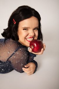 The author, Jillian, is wearing a mesh black top with shining silver constellations all over. Under it is a majestically black strap cami top. Their hair is done in the fashion of Snow White with a red hair clip and they are smiling at the viewer while holding an apple in their right hand and lying down. They have a stunning smile with red lipstick.