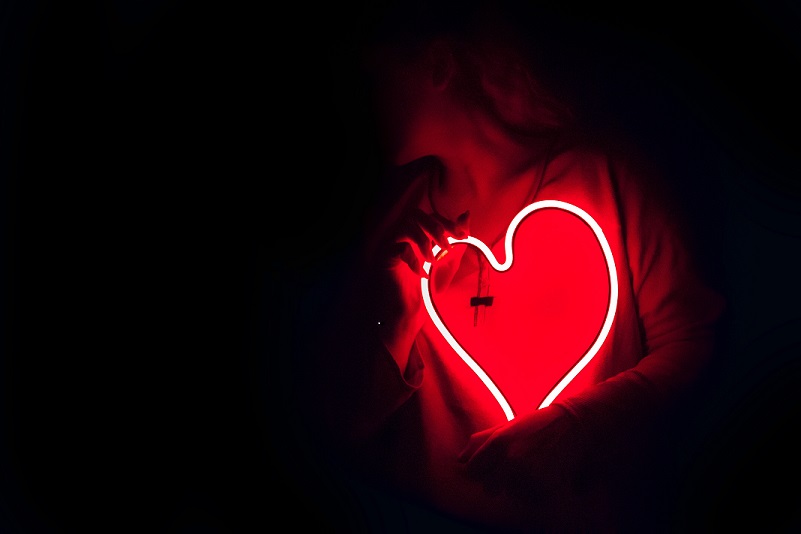A red glowing heart held by a person in a dark room. They are holding it on their chest.