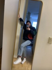 Kathryn is pictured in front of a long mirror. She is wearing an oversized sweater, straight-cut jeans, floral socks, and high-top white Converse. She is posed with her hand in the air and foot off the ground to display her outfit clearly.